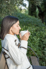 Young girl drinking from a cup in the field. Sweater for cold autumn or winter. Breathe and smell the aroma of coffee. Vertical