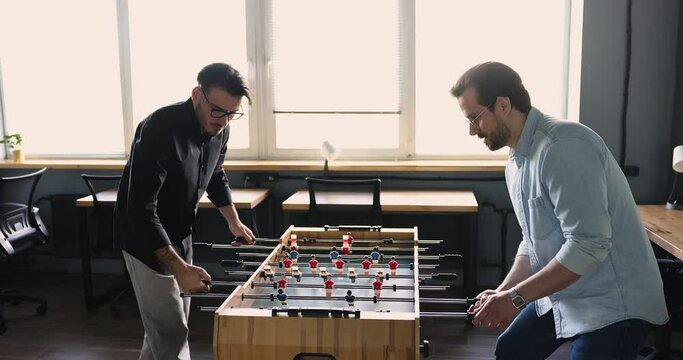 Two handsome 30s businessmen have fun playing together foosball in office, winning match, cheering looking overjoyed, spending stress-free break at workplace, engaged in hobby, enjoy favourite game