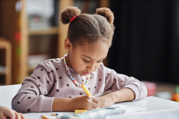 Cute serious African American schoolgirl drawing with yellow highlighter while sitting by table at lesson of craft and art in kindergarten - obrazy, fototapety, plakaty