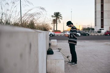 caucasian guy standing in profile with piercing in his nose striped sweater looking at camera the screen of his smartphone in his hand next to the skateboard in the city