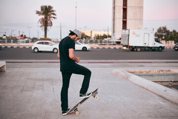 caucasian young man in black t-shirt and cap on his head standing on his skateboard in the city