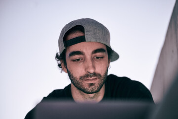 face of bearded caucasian guy with nose piercing gray cap black t-shirt sitting looking at his laptop screen on a city street