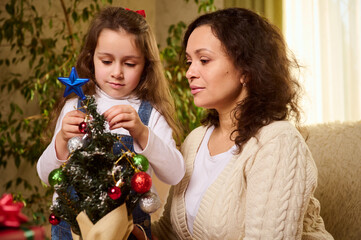 Beautiful multi-ethnic woman, a loving mother in warm beige sweater, decorates an eco Christmas tree with her adorable lovely daughter, a little girl hanging toys on Xmas tree in cozy home interior