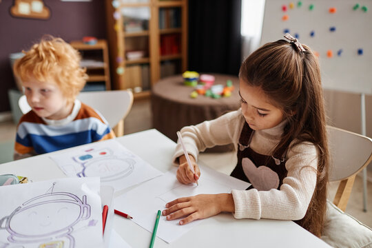 Little learner of nursery school drawing picture with crayons on paper while sitting by desk against gingerhead schoolboy in classroom