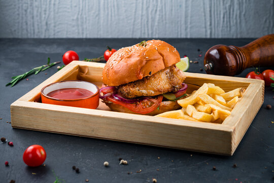 Chicken burger with tomato, pickle, onion, lettuce, cheese sauce on a wooden board with french fries and ketchup.