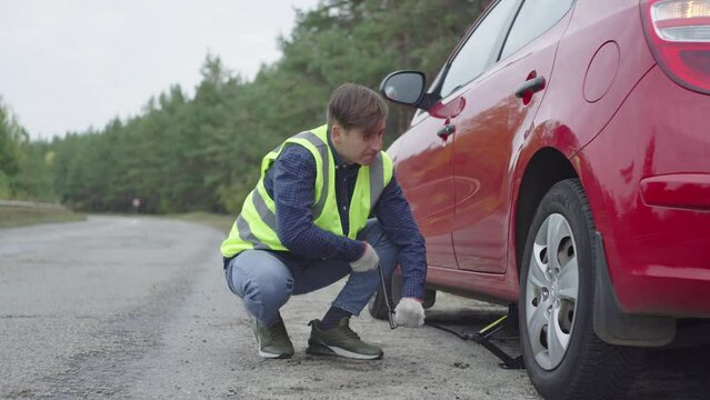 A man in a vest installs a jack to change a tire in a car. Car on the side of the road. Replacement of a damaged wheel. Road accident on the highway. The guy hangs up the car. High quality 4k footage