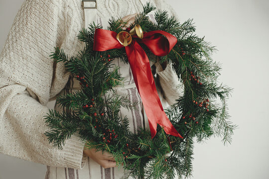 Woman holding stylish christmas wreath with red ribbon and golden bells close up on white wall background. Making Christmas wreath, moody image. Winter holiday decor. Merry Christmas!