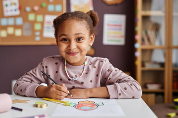 Cute successful learner of primary school looking at camera with smile while sitting by table and drawing with crayons at lesson - obrazy, fototapety, plakaty