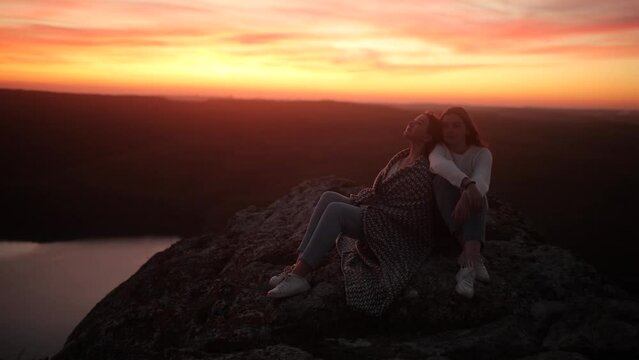 Two teenage girls, wrapped in blankets, sit on top of a rock and enjoy the summer sunset. Sisters enjoy youth and freedom. A gust of wind blows the hair of two friends