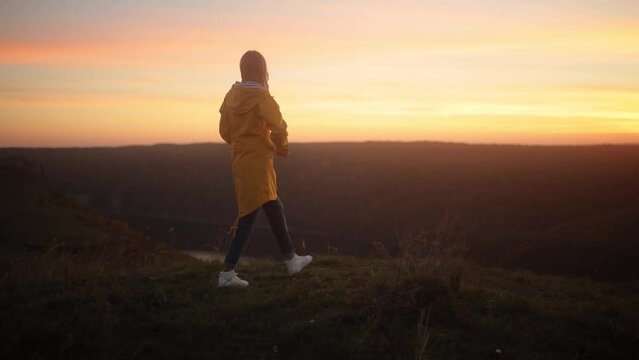 Camera follows young woman in yellow jacket running up on top of mountain summit at sunset, jumps on top of rocks, raises arms into air, happy and drunk on life, youth and happiness