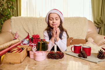 Delightful kid, a baby girl in Santa hat, keeps hands palms together, makes a cherished wish,...