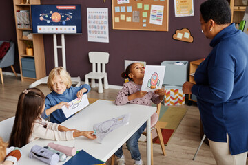 Cute little learner of primary school boasting with her drawing of rocket while showing paper to African American teacher at lesson