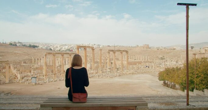 Young woman sitting on bench in front of North Colonnaded Street in Gerasa, Jerash, Jordan. Brown haired lady looking at ancient ruins and landscape with architecture. 4k zoom in shot
