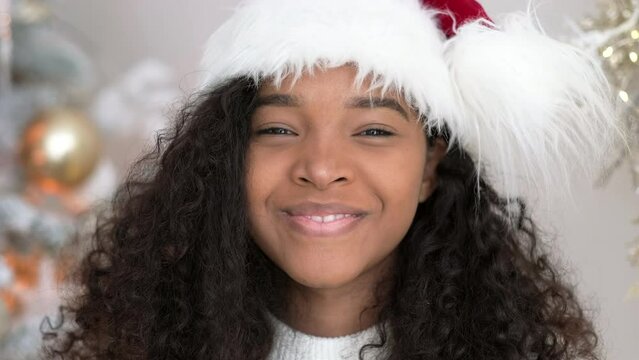 Christmas and New Year portrait beautiful smiling African American girl in Santa hat near Christmas tree, Smiling young black woman with curly hair looking at camera.