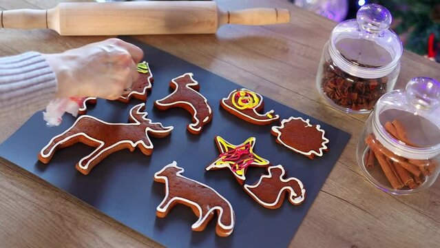 Women's hands coloring Christmas gingerbread cookies at home in slow motion. Christmas decoration in the kitchen. Fir tree with fairy lights. The concept of the New Year and Christmas