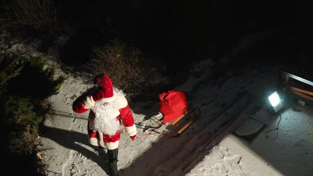 Santa Clause pulling sledge walking from darkness at night putting red bag with presents on snowy porch. High angle view wide shot of Caucasian man outdoors on Christmas night. Miracles and surprise