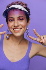 Portrait of a sporty fashion woman posing smiling with teeth at the camera with stylish earrings in her ears in a purple yoga tracksuit and a transparent cap on a purple monochrome background