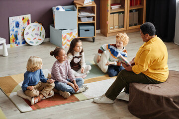 Several adorable intercultural kids gathered on the floor in front of their teacher of nursery school reading them book of tales