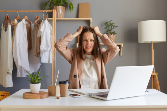Image of aggressive angry Caucasian brown-haired woman stylist wearing beige jacket sitting at her workplace in front of notebook, screaming, keeping hands on head.