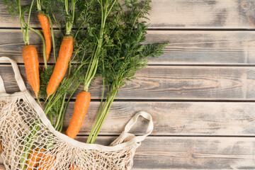 Orange tasty juicy carrot with green stem and leaves in ecological string bag on wooden background....