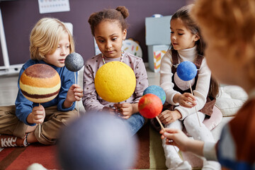 Cute little learners of primary school with models of solar system planets discussing their...