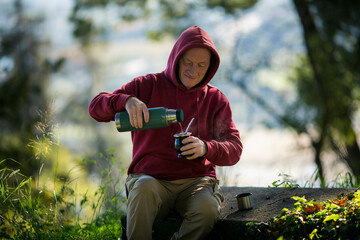 A man in the park pours boiling water from a thermos into a Mate cup. - 547790896