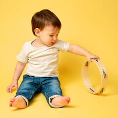 Toddler baby plays the tambourine, a child with a percussion musical instrument on a studio yellow background. Happy child musician playing hand drum. Kid aged one year four months - obrazy, fototapety, plakaty