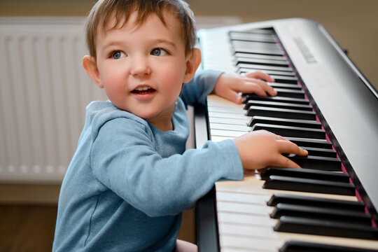 Toddler baby plays piano in home living room, music lessons. Happy child boy learns to play music on an electric piano. Kid boy age one year four months