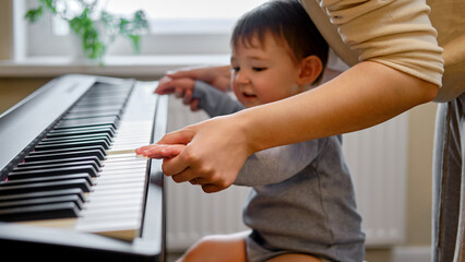 A mother woman teaches a toddler baby to play the piano in a home living room. An adult teacher...