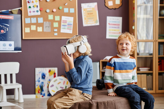 Little boy in vr headset watching video and playing virtual game while sitting next to happy classmate in classroom at nursery school