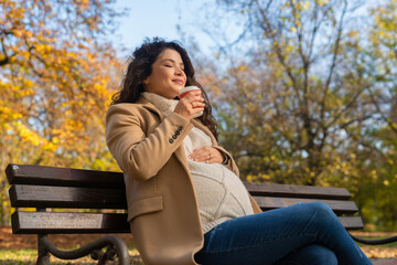 Pregnant woman sits on bench in the park drinking coffee