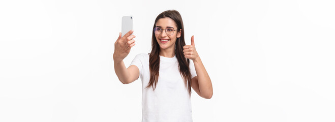 Fototapeta na wymiar Communication, technology and lifestyle concept. Portrait of upbeat happy, smiling caucasian girl in glasses, internet blogger taking selfie, record video message to followers thumbs-up, hold phone