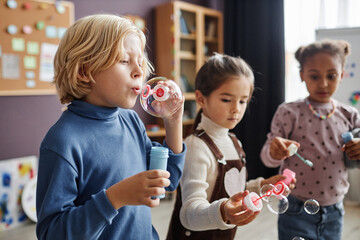 Cute blond boy blowing soap bubbles in front of camera against two intercultural girls while having fun in nursery school - obrazy, fototapety, plakaty