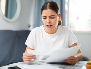 Disappointed young adult woman sitting at table at home, reading documents, having bad news