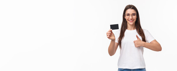 Fototapeta na wymiar Portrait of pleased, attractive young brunette woman in t-shirt and glasses, recommend bank, showing credit card, smiling satisfied show thumbs-up, approve like and advice make online purchases