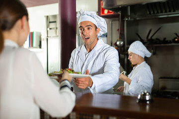 Fototapeta na wymiar Portrait of smiling man chef working in open restaurant kitchen, giving out ready meals to waitress on order station