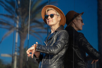 Tourist 30s blond woman with short hair in classy hat, sunglasses and leather jacket with backpack next to modern mirror building in Barcelona