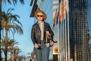 30s blond woman with short hair in classy hat, sunglasses and leather jacket next to modern building in Barcelona with european flags on background