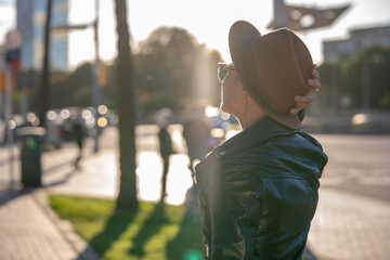 30s blond woman with short hair in classy hat, sunglasses and leather jacket next to modern building in Barcelona with back sunlight