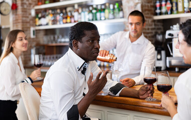 Multiracial couple drinking wine in loft-style bar with young bartender serving them and a single woman on background - obrazy, fototapety, plakaty