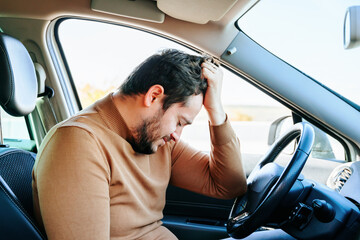 A young man is in a car sitting with his elbow on the steering wheel and his hand dug into his...