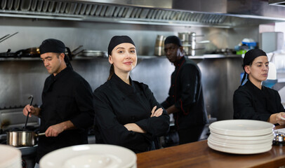 Portrait of smiling woman head chef posing in modern restaurant kitchen, proud of good work of her team of chefs