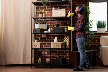 Housewife wearing protection gloves dusting furniture shelves in modern apartment, while listening music using headphones. Tired woman cleaning new home in the city they just moved into.