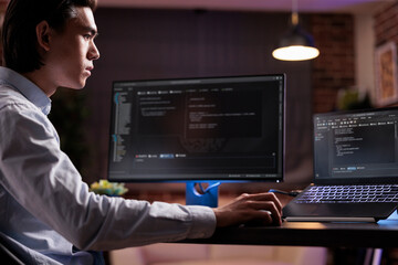 Software developer coding firewall program on monitor and laptop, using encryption server to script...
