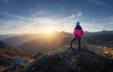 Fototapeta premium Young woman on mountain peak and beautiful mountains in haze at colorful sunset in autumn. Dolomites, Italy. Sporty girl, mountain ridges in fog, orange grass, trees, golden sun in fall. Hiking