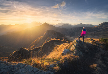 Young woman on mountain peak and beautiful mountains in haze at colorful sunset in autumn. Dolomites, Italy. Sporty girl, mountain ridges in fog, orange grass, trees, golden sun in fall. Hiking	 - obrazy, fototapety, plakaty