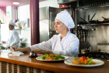 Portrait of young adult female chef cook checking orders and handing plate with meal at restaurant kitchen