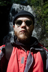 male traveler with a beard on a hike against the backdrop of a waterfall in the mountains