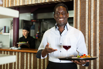 Portrait of confident man waiter holding plate of salad and glass of wine in restaurant with open...