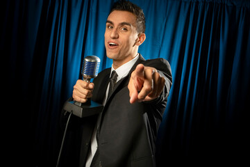 Man dressed in a suit and tie pointing at the camera talking into a microphone on a blue curtain...